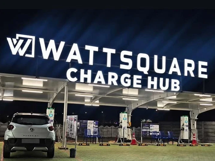 Wattsquare Charge Hub at night: illuminated signage over a steel canopy with DC chargers and a car charging in the bay